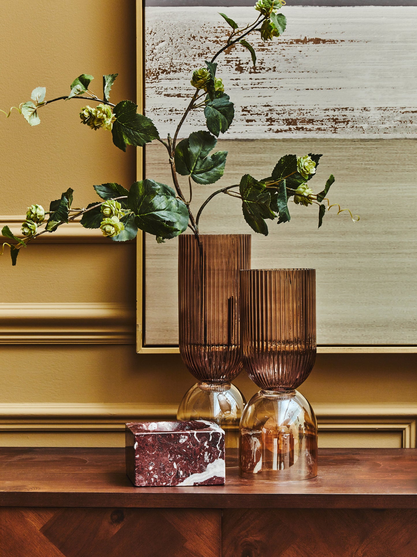 Two brown glass vases with a marble candle holder on a wooden surface, against a gold wall with a framed abstract painting.